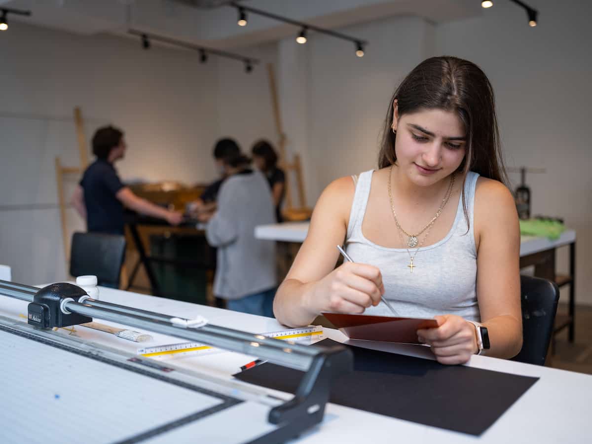 Estudiante de la Universidad Anáhuac trabajando en un proyecto de diseño en su mesa de trabajo, utilizando herramientas como una regla y cuchilla en un entorno creativo.
